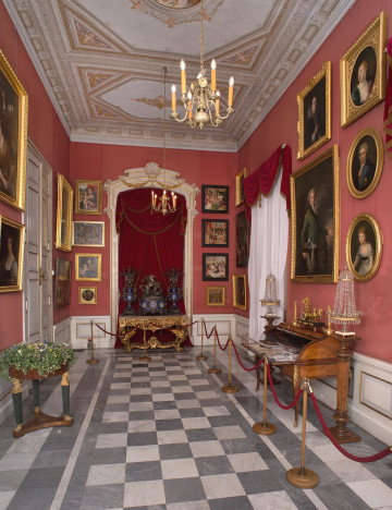 ściana południowa, w tle portal drzwiowy ozdobiony udrapowaną bordowa tkaniną, na różowych ścianach wiszą obrazy, po prawej sekretarzyk i okno z białą firaną i czerwonym lambrekinem, podłoga z szaro-białe kwadraty w kształcie szachownicy.