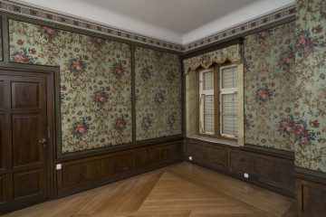 malarsko-sztukatorska dekoracja ściany W 67M0009, narożnik północno-wschodni, ściany obite tkaniną, od dołu cokół na wzór boazerii, po lewej zamknięte drzwi drewniane, po prawej okno z zamkniętymi zewnętrznymi okiennicami.
