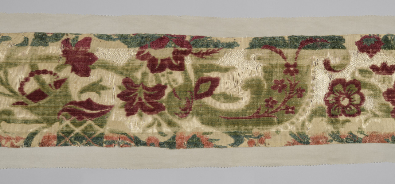 Pas tkaniny ułożony poziomo – zbliżenie na fragment. Widoczne motywy roślinne: czerwone kwiaty poprzeplatane zielonymi liśćmi na beżowym tle. W centralnej części szeroki pas tkaniny jest odbarwiony, liście są jaśniejsze, w tle jaśniejsze przeszycia. Wąski pas na dole również odbarwiony, motywy kwiatowe wpadają w pomarańcz. bryt Wil.Wn.126/1 - fragment, awers; ściana północno-wschodnia