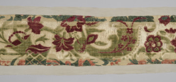 Pas tkaniny ułożony poziomo – zbliżenie na fragment. Widoczne motywy roślinne: czerwone kwiaty poprzeplatane zielonymi liśćmi na beżowym tle. W centralnej części szeroki pas tkaniny jest odbarwiony, liście są jaśniejsze, w tle jaśniejsze przeszycia. Wąski pas na dole również odbarwiony, motywy kwiatowe wpadają w pomarańcz. bryt Wil.Wn.126/1 - fragment, awers; ściana północno-wschodnia