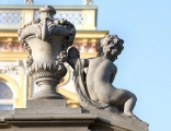 ujęcie od strony trzeciej. Putto z prawej, wazon z lewej. Putto ustawione na pochyłej balustradzie schodów, wazon na poziomej. Putto ma tułów silnie skręcony w lewą stronę i jest zwrócone plecami do widza. Draperia przewieszona przez lewe ramię. W rękach wyciągniętych w lewo trzyma kwiatową girlandę. Głowa zwrócona w prawo. Obok waza o kielichowatym kształcie dekorowana girlandami kwiatów i stylizowanymi liśćmi. Obie figury w kolorze beżowym. W tle fasada pałacu.
