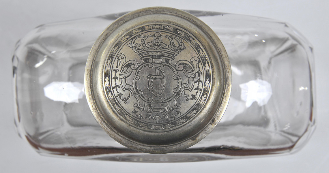 Ujęcie z góry - zakrętka z grawerowanym herbem Janina - wygięta tarcza skierowana w prawo. Nad herbem korona królewska, a całość otoczona wieńcem. Na drugim planie widoczna szeroka, płaska butla o płaskich i przezroczystych ściankach.