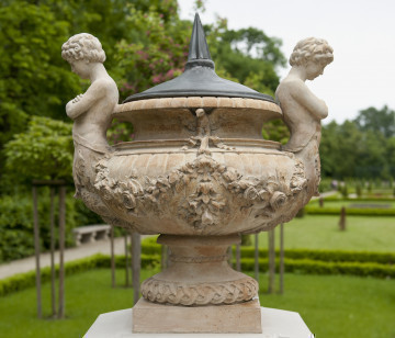 ujęcie od strony pierwszej. Wazon dekoracyjny na wysokim trzonie, szeroki z wywiniętą krawędzią. Ustawiony na wysokiej podstawie. Kolor beżowy. Dookoła brzuśca kwiatowa girlanda. Dwa symetryczne uchwyty w kształcie młodzieńców z podwójnymi rybimi ogonami – po lewej i prawej stronie. Przy szyjce ptak z rozpostartymi skrzydłami. Wazon przykryty metalowym hełmem ze szpiczastym uchwytem. W tle widoczny park.