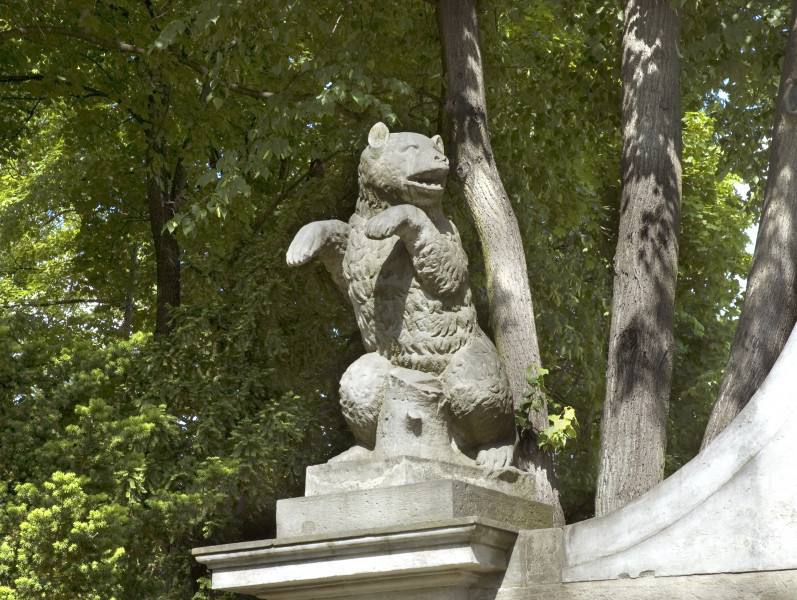 Niedźwiedź - rzeźba na przyporze północnej Bramy Głównej. Widok lekko z prawej i od dołu. Niedźwiedź stoi na dwóch tylnych łapach. Dwie przednie łapy wyciągnięte przed siebie. Łeb niedźwiedzia skierowany w prawo. Między tylnymi łapami widoczny pień drzewa.W tle zielone liście i pnie drzewa.