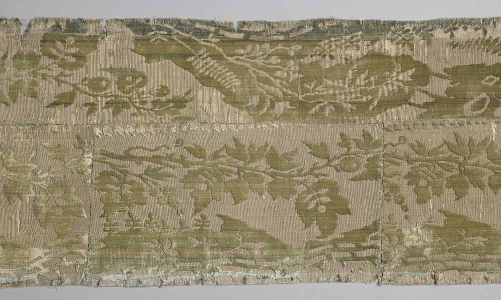Pas tkaniny z motywami roślinnymi w kolorze żółto-zielonym. Na górze pas odbarwiony na niebiesko. Widoczny poziomy szew oraz dwa pionowe szwy sięgające od dołu do poziomego szwu. bryt Wil.Wn.132/1 - fragment, awers; ściana północna