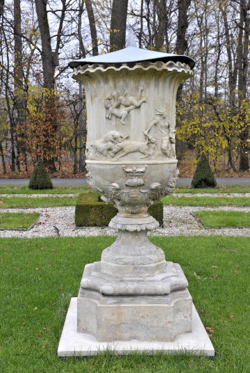 ujęcie od strony czwartej, widok z boku, wazon przykryty metalową nakrywką z ostrym trzpieniem, czasza wazonu ozdobiona sceną lecących amorków siłujących się, oraz zwierząt podczas polowania,  dół wazonu ozdobiony tarczami herbowymi, a noga wazonu ozdobiona wzorami kwiatowymi, wazon stoi na cokole wśród trawy w tle stożki cisów oraz drzewa bez liści.