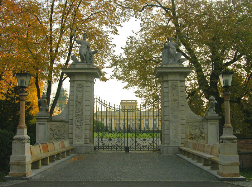 Widok na Bramę Główną od strony Przedpola, przęsła metalowe zamknięte. Brama w kolorze beżowym, o bogato zdobionej elewacji. Rzeźby mają kolor szary. Po lewej i prawej na przyporach rzeźby kolejno niedźwiedzia i psa. Łączące się niewielkim łukiem z wysokimi filarami. Na filarach widoczne rzeźby. Na lewym – Mars, a na prawym – Pax. W tle widoczny korpus Pałacu. Do bramy prowadzi brukowany, krótki most nad fosą. Wzdłuż jego lewej i prawej strony stoją w rzędzie kamienne ławy. Na początku każdego rzędu stoi kamienna, kolumnowa latarnia. Za bramą drzewa, pełne jesiennych liści.