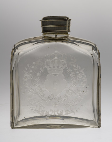 Ujęcie z przodu, na całość butli. Ścianka szeroka, płaska, bezbarwna. Widoczny wygrawerowany na niej herb Janina – wygięta tarcza skierowana w prawo. Herb otoczony zdobieniami. Ścianka na szczycie łagodnie się zwęża do srebrnego obramienia szyjki. Na butlę nałożona jest nakrętka. Przez szkło widoczne dekoracje na przeciwległej ściance.