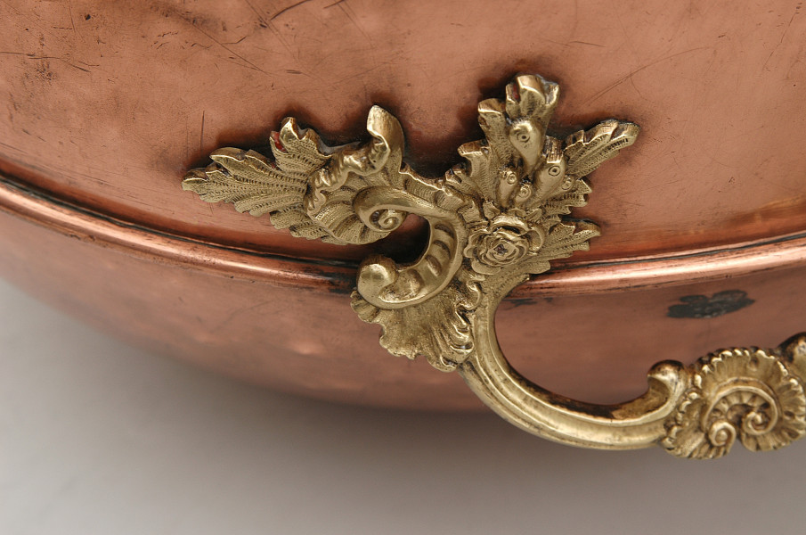 Fragment cyzelowanego ucha z ornamentem roślinnym (stylizowany kwiat róży i liście) na tylnej ściance. Widoczna część lewa uchwytu. Ucho w kolorze złotym. Ścianka pojemnika w kolorze jasno-brązowym.