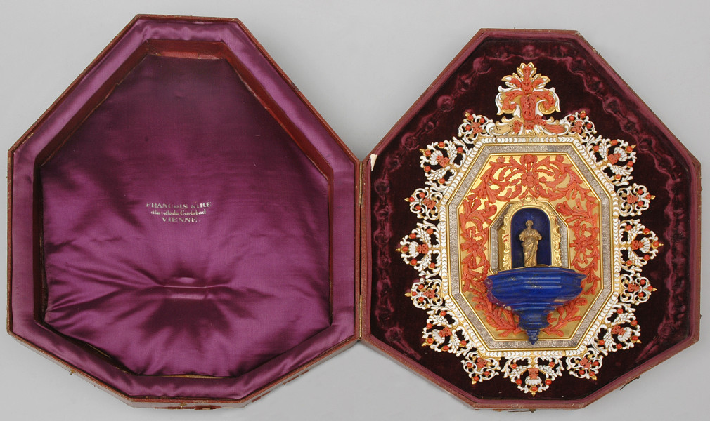 kropielniczka na ośmiokątnej tarczy z emalowaną ramką w futerale.Po prawej stronie w pokrowcu kropielniczka bogato dekorowana, otoczona ażurową biało emaliowaną ramką z koralami nad niebieską kropielniczką, we wnęce Madonna z brązu złoconego. Pokrowiec ośmiokątny, wewnątrz wyłożony fioletowym atłasem i aksamitem, po lewej stronie wytłoczone Francois Siré a la ville de Carlsbad VIENNE.
