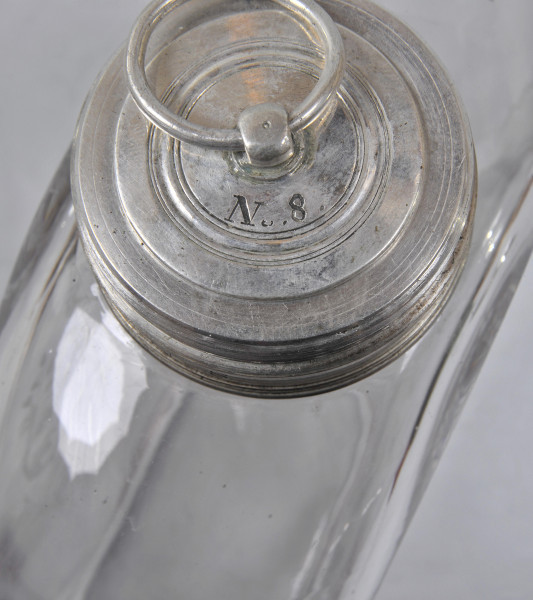 Ujęcie z góry, zbliżenie na zatyczkę. Zatyczka okrągła, srebrna. Na wierzchu grawerowane koncentryczne okręgi, wewnątrz nich wygrawerowany napis „N.6.”. W górnej części kadru uchwyt w formie niewielkiej obręczy, przewleczony przez kulistą wypukłość na środku. Uchwyt nie mieści się w kadrze. Na drugim planie widoczna szeroka, płaska butla o płaskich i przezroczystych ściankach. Butelka ustawiona jest pod kątem.