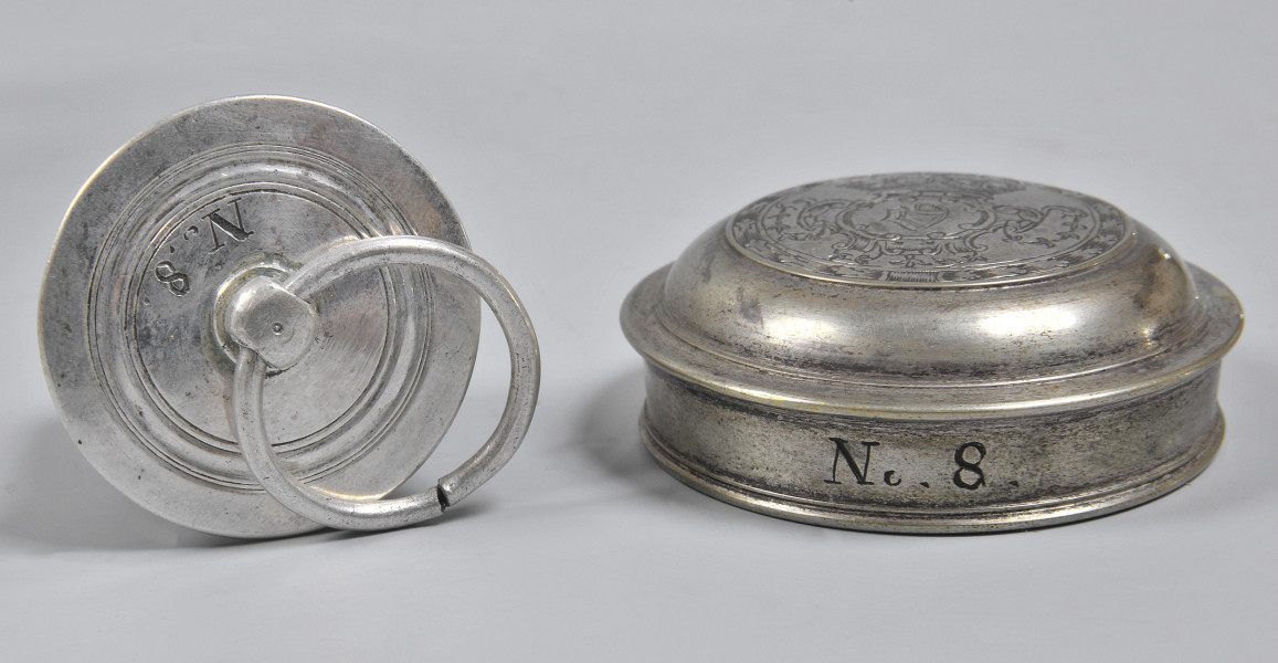 Zatyczka-korek i nakrętka. Zatyczka okrągła, srebrna, po lewej stronie, ustawiona bokiem. Na wierzchu grawerowane koncentryczne okręgi, wewnątrz nich wygrawerowany napis „N.6.”. O ziemie oparty jest uchwyt w formie niewielkiej obręczy, przewleczony przez kulistą wypukłość na środku. Po prawej srebrna nakrętka. Na jej boku wygrawerowany napis „N.8.”. Na wierzchu widoczne wygrawerowane dekoracje.