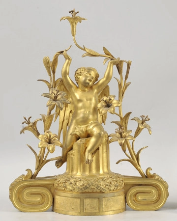 ujęcie z przodu. Złota w kolorze figura skrzydlatego chłopca. Chłopiec siedzi zwrócony w prawo. Głowę ma odchyloną do tyłu i skręconą w prawo. W rękach uniesionych nad głową podtrzymuje wysokie pędy lilii.