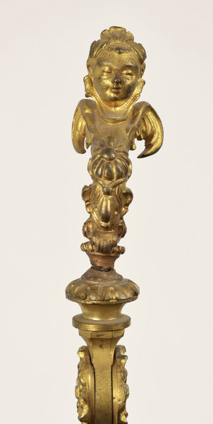 Detal rękojeści - ujęcie przodu. Rękojeść bogato zdobiona w kolorze złotym. Na szczycie znajduje się figurka hermy o dziecięcej głowie.