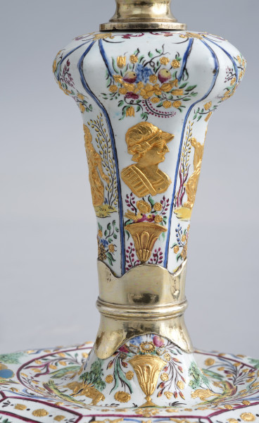 zbliżenie na dekorację nóżki lichtarzyka, na dole fragment podstawy ze złotym dzbanem z kwiatami, powyżej złoty pierścień w centralnej części popiersie mężczyzny w hełmie pod nim złoty dzban z kwiatami a nad nim girlanda z kwiatów i owoców. U góry złoty element.