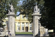 Widok na Bramę Główną od strony Przedpola, w tle korpus pałacu. Po lewej i prawej widoczne przypory łączące się niewielkim łukiem z wysokimi filarami. Na filarach widoczne rzeźby. Na lewym – Mars, a na prawym – Pax. Przęsła metalowe otwarte. Po lewej i prawej na pierwszym planie drzewa.