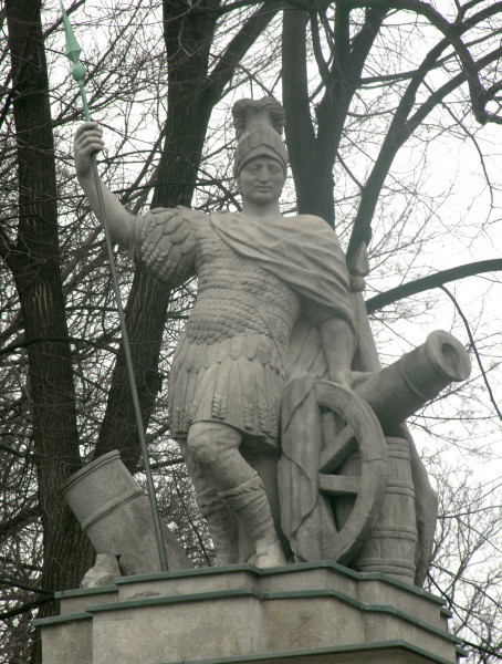 Mars - rzeźba na filarze północnym Bramy Głównej. Ujęcie lekko z prawej i z dołu. Mężczyzna ubrany w zbroję łuskową i płaszcz okrywający lewą rękę. Na głowie założony rzymski hełm z pióropuszem. Lewa dłoń oparta na skierowanej w prawo armacie. W prawej dłoni postać trzyma skierowaną w górę włócznię. Twarz skierowana w kierunku widza, nieco powyżej kadru. W tle gałęzie drzew.