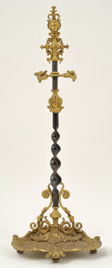 Ujęcie z przodu. Na dole zdobiona podstawka z widocznymi trzema nóżkami wystającymi od spodu. Na podstawce osadzony trzpień do połowy wysokości skręcony w spiralę. Powyżej na trzpień nałożone zostały dwie przewiązki. U góry trzpień zdobi płaska sterczyna z motywem małżowinowym i z płaskorzeźbą kobiecej twarzy. Trzpień w kolorze czarnym, pozostała cześć obiektu złota.