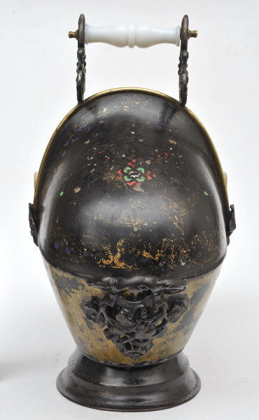 Ujęcie z tyłu. Pojemnik o dzwonowatym kształcie osadzony na czarnej stopce. Na pierwszym planie widoczny czarny, tylny uchwyt z motywem roślinnym. Na drugim planie, przy krawędzi pojemnika, uchwyt górny z podwójnych zwojów i okładką z białego, mlecznego szkła. Na pojemniku widoczne wytarte dekoracje malarskie. Pojemnik w kolorze czarnym z żółtymi wytarciami.