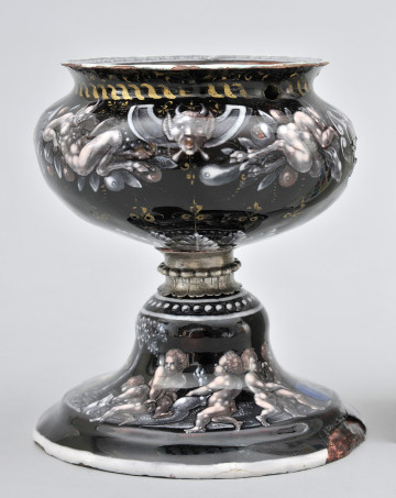 ujęcie z boku. Górna część czasza, na czarnym tle biało-szara dekoracja, głowa maszkarona w centralnej części, po bokach  całe postacie maszkaronów na tle kiści owoców. Srebrny łącznik z nóżką. Dolna część stopka ozdobiona dekoracją leżący niedźwiedź na nim putta.
