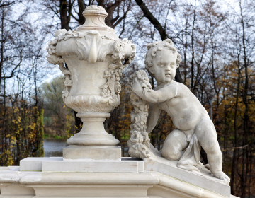 ujęcie od strony pierwszej. Putto z prawej, wazon z lewej. Putto ustawione na pochyłej balustradzie schodów, wazon na poziomej. Putto oparte na prawym kolanie, lewa noga wyprostowana. Prawa ręka wyprostowana wsparta na podstawie, w lewej ręce girlanda, tułów silnie skręcony w prawą stronę. Chłopiec zwrócony twarzą w stronę widza. Obok waza o kielichowatym kształcie dekorowana girlandami kwiatów i stylizowanymi liśćmi. Obie figury w kolorze beżowym. W tle drzewa parkowe.