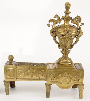 ujęcie z przodu (strona pierwsza). Złoty w kolorze wilk napięciu zbieżnych nóżkach, o masywnej części poziomej, złożonej z dwóch prostopadłościanów. Po prawej stronie widoczny szerszy, stanowiący cokół dla bogato rzeźbionej sterczyny w formie wazy zwieńczonej płomieniem. Po lewej węższy, stanowiący podporę dla polan.