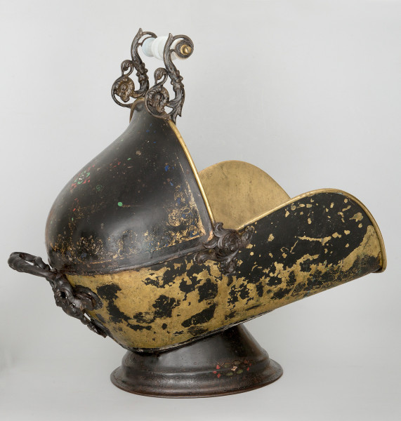 Ujęcie 3/4. Pojemnik widoczny z boku i lekko z tyłu. Umieszczony na stopce, pochylony lekko w gorę. Kształt przypominający dzwon, z wyciętą górną prawą ćwiartką. W rogu wycięcia umieszczona dekoracja z motywem roślinnym. Widoczne są dwie rączki: górna, na krawędzi pojemnika, i tylna, z lewej strony. Pojemnik czarny, kolor żółty w obdrapanych miejscach. Większość obdrapań w dolnej części.