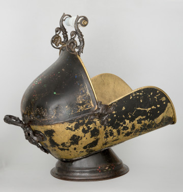 Ujęcie 3/4. Pojemnik widoczny z boku i lekko z tyłu. Umieszczony na stopce, pochylony lekko w gorę. Kształt przypominający dzwon, z wyciętą górną prawą ćwiartką. W rogu wycięcia umieszczona dekoracja z motywem roślinnym. Widoczne są dwie rączki: górna, na krawędzi pojemnika, i tylna, z lewej strony. Pojemnik czarny, kolor żółty w obdrapanych miejscach. Większość obdrapań w dolnej części.