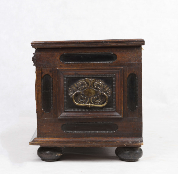 Korpus prostokątny, zamykany od góry gładkim  wiekiem na zawiasach, cztery nogi w kształcie silnie spłaszczonych kul. Ściana frontowa podzielona pionowo na dwie części, w których umieszczone są poziome płyciny w kształcie prostokąta o ściętych narożach wypełnionych trójkątnymi rautami, ujęte w profilowane ramy. Podziałowi odpowiadają w górnej części trzy rzeźbione maski lwów. Wzdłuż boków ściany frontowej i bocznych - lekko wgłębione płyciny w formie wydłużonych owali. Wszystkie płyciny i rauty obwiedzione ornamentem wody. W ścianach bocznych zamocowane masywne metalowe antaby.  Wewnątrz żelazny zamek nakładany.    