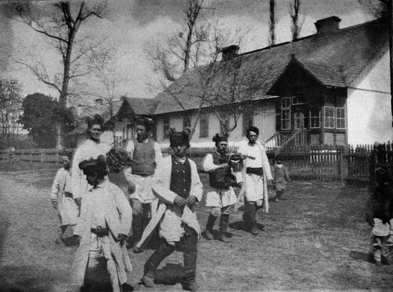 Fotografia czarno-biała przedstawia orkiestrę weselną z instrumentami muzycznymi. W oddali widoczne drewniane zabudowania wiejskie.