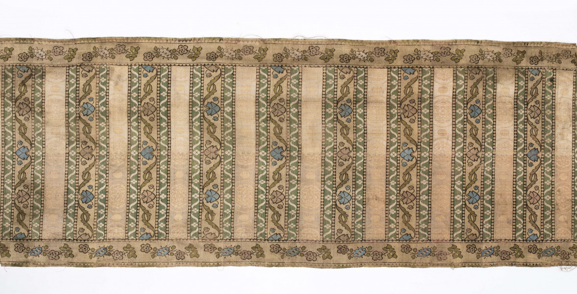 Pas 4-stronny. 
Ornament: głowy - krzaczki kwiatowe w wazonach; środek - poprzeczne pólka w układzie a,b,a,c (a - motyw wijacego sie wężyka, b - medaliony z rozetami, c - wić kwiatowa), obrzeża pionowe z gałązkami kwiatowymi, poziome - jak pólka c.
Koloryt: prawa strona - tło słomkowe, nieznacznie zróżnicowane w każdej połowie, zielen, błękit, żółty; lewa strona - tło brązowe głów i obrzeżeń, ornament - tło słomkowe, wzór j.w..
Fredzla doszyta jedwabna, pleciona, słomkowożółta