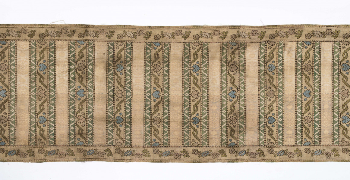 Pas 4-stronny. 
Ornament: głowy - krzaczki kwiatowe w wazonach; środek - poprzeczne pólka w układzie a,b,a,c (a - motyw wijacego sie wężyka, b - medaliony z rozetami, c - wić kwiatowa), obrzeża pionowe z gałązkami kwiatowymi, poziome - jak pólka c.
Koloryt: prawa strona - tło słomkowe, nieznacznie zróżnicowane w każdej połowie, zielen, błękit, żółty; lewa strona - tło brązowe głów i obrzeżeń, ornament - tło słomkowe, wzór j.w..
Fredzla doszyta jedwabna, pleciona, słomkowożółta