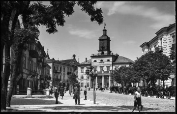 Fotografia czarnobiała z widokiem Nowego Ratusza w Lublinie od strony ulicy Królewskiej. 
Widok zamykający perspektywę ulicy Królewskiej. W centralnej części klasycystyczny budynek z czterokolumnowym portalem i wieżą zwieńczoną herbem miasta Lublina z wizerunkiem koziołka. Nad wejściem napis „Magistrat”. W głębi po lewej kopuła kościoła p. w. Św. Ducha oraz kamienice przy ulicy Krakowskie Przedmieście. Po prawej, przed kamienicą rząd rozłożystych drzew, pod którymi stoją zaparkowane dorożki konne. Po lewej fasady kamienic z szyldami. Na szerokim chodniku, oddzielającym od brukowanej ulicy, przechodnie. 