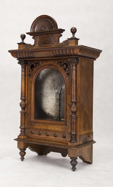 Pozytywka w szafce eklektycznej, drewnianej pokrytej okleiną z drewna orzechowego. Szafka prostokątna, pionowa, z ozdobnym zwieńczeniem, na niskich nóżkach, na froncie drzwiczki. Pośrodku zwieńczenia prostokątny cokolik z muszlą, po bokach kuliste sterczynki na sześciennych podstawach. Gzyms wieńczący górą wysunięty z rytowanym pośrodku  napisem 