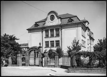 Fotografia czarnobiała gmachu Gimnazjum im. Stefana Batorego w Lublinie (tzw. Szkoły Lubelskiej) przy ul. Spokojnej. 
Widoczny front budynku zamykającego perspektywę ulicy Ewangelickiej oraz fragment jednej ze ścian bocznych. Gmach wolnostojący, otynkowany, dwukondygnacyjny, kryty spadzistym mansardowym dachem ze szczytami i lukarnami po brzegach. Budowla o cechach architektury secesyjnej. Pośrodku widocznej od ulicy elewacji dwa rzędy prostokątnych okien, po trzy na każdym poziomie. Nad nimi, na poddaszu, w centralnej części owalne okno. Teren szkoły oddzielony od ulicy ogrodzeniem z metalowych pionowych, poziomych i giętych prętów przedzielonych murowanymi prostokątnymi filarami. Na szczytach słupków ozdobne elementy (m. in. w kształcie kuli). Brama wjazdowa łukowato zwieńczona. Po obu jej stronach symetrycznie rozmieszczone furtki. Przed wejściem dwie tablice urzędowe z numeracją budynku. Wokół dużo zieleni. W prawym dolnym rogu kadru dekoracyjnie przystrzyżony żywopłot. 