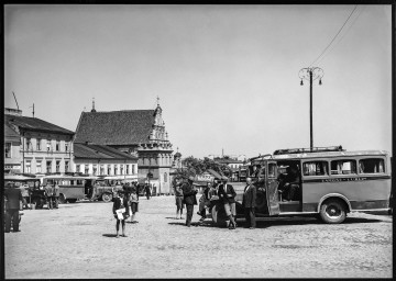 Fotografia czarnobiała dworca autobusowego przy ul. Świętoduskiej w Lublinie z kościołem Karmelitów w tle oraz autobusem linii Zamość-Lublin. Z prawej strony kadru zaparkowany autobus zamiejscowy linii Zamość-Lublin. Kilku mężczyzn stoi opartych o przednią maskę. Drzwi pojazdu są uchylone. Wewnątrz siedzą dwie osoby. Na bocznej karoserii naniesiona informacja z docelowymi punktami kursu. Za autobusem wysoka uliczna latarnia. Pośrodku bosy chłopiec w czapce z daszkiem trzyma w ręku złożoną gazetę (widoczny tytuł „ABC”). Obok niego dwaj chłopcy w wieku szkolnym. W tle po lewej widoczna bryła kościoła Karmelitów i przyległe do niej po obu stronach pierzeje kamienic. Przed zabudowaniami zaparkowane autobusy oraz pasażerowie.