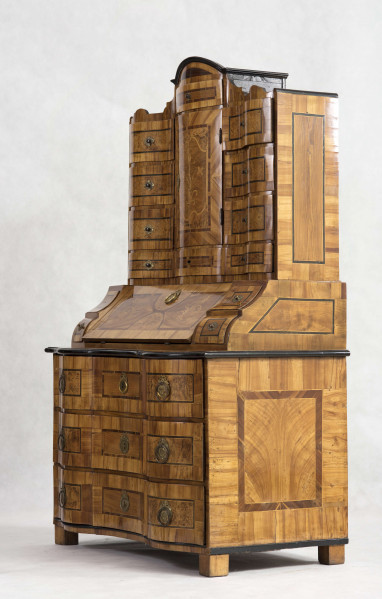 Sekretera trójkondygnacyjna. Podstawa w formie trzyszufladowej komody,wspartej na czterech kostkowych nogach. Środkowa część z prostokątnym skośnym, odchylnym blatem, obwiedziona profilowaną listwą, fronty szuflad wklęsło-wypukłe. Po bokach ukośnego blatu po dwie szufladki. Nastawa cofnięta z dwoma rzędami szufladek po bokach (po 4); część środkowa wyższa, w niej półkoliście wypukłe drzwiczki i dwie szufladki nad i pod drzwiczkami. Fronty szuflad faliste jak i wykrój profilowanego zwieńczenia. Powierzchnia intarsjowana, dekorowana fornirem: na drzwiczkach kompozycja w formie gałązki z kwiatami, na blacie symetrycznie ułożone dwa ornamenty liściasto-wolutowe,  na szufladach poziome prostokątny ujęte w ciemne ramki. Nakładane brązy:  szyldziki do kluczy, antabki i kuliste uchwyty. 
