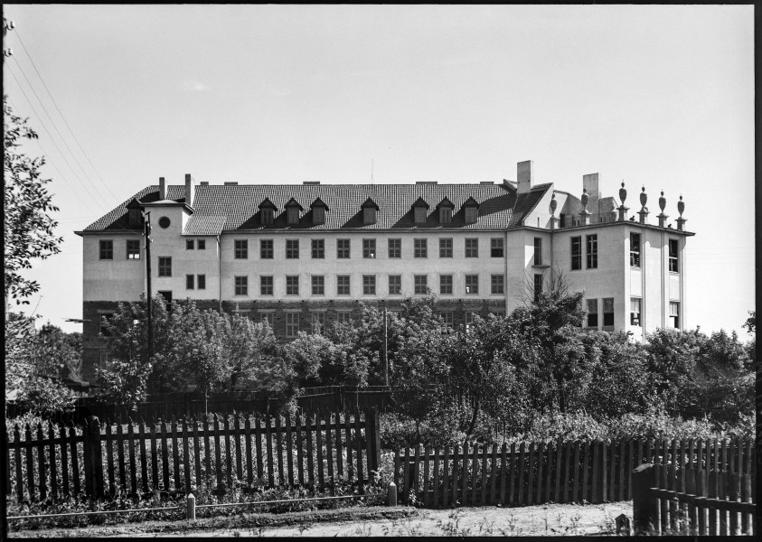 Fotografia czarnobiała przedstawiająca Wyższe Gimnazjum Biskupie Męskie („Biskupiak”, obecne XXI LO im. św. Stanisława Kostki w Lublinie przy ul. Słowikowskiej) od strony ul. Szkolnej (prawdopodobnie wykonana z budynku obecnego IV Liceum Ogólnokształcącego). Na pierwszym planie widoczny drewniany płot ogradzający teren zielony z gęsto posadzonymi drzewami. W oddali modernistyczny gmach szkolny. Widoczne skrzydło zachodnie oraz ryzalit południowy zwieńczony ośmioma ozdobnymi postumentami z imitacją obecnie nie istniejących amfor zwieńczonych krzyżykami. Pierwsze piętro budynku z odsłoniętym wątkiem ceglanym, drugie otynkowane i pobielone; dach pokryty dachówką.