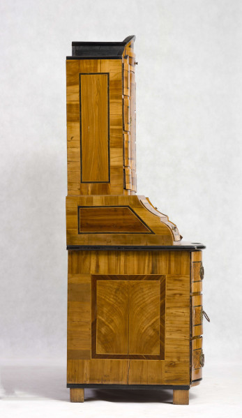 Sekretera trójkondygnacyjna. Podstawa w formie trzyszufladowej komody,wspartej na czterech kostkowych nogach. Środkowa część z prostokątnym skośnym, odchylnym blatem, obwiedziona profilowaną listwą, fronty szuflad wklęsło-wypukłe. Po bokach ukośnego blatu po dwie szufladki. Nastawa cofnięta z dwoma rzędami szufladek po bokach (po 4); część środkowa wyższa, w niej półkoliście wypukłe drzwiczki i dwie szufladki nad i pod drzwiczkami. Fronty szuflad faliste jak i wykrój profilowanego zwieńczenia. Powierzchnia intarsjowana, dekorowana fornirem: na drzwiczkach kompozycja w formie gałązki z kwiatami, na blacie symetrycznie ułożone dwa ornamenty liściasto-wolutowe,  na szufladach poziome prostokątny ujęte w ciemne ramki. Nakładane brązy:  szyldziki do kluczy, antabki i kuliste uchwyty. 
