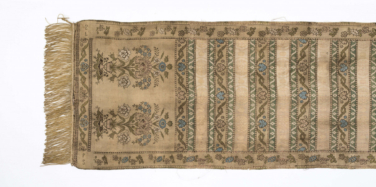 Pas 4-stronny. 
Ornament: głowy - krzaczki kwiatowe w wazonach; środek - poprzeczne pólka w układzie a,b,a,c (a - motyw wijacego sie wężyka, b - medaliony z rozetami, c - wić kwiatowa), obrzeża pionowe z gałązkami kwiatowymi, poziome - jak pólka c.
Koloryt: prawa strona - tło słomkowe, nieznacznie zróżnicowane w każdej połowie, zielen, błękit, żółty; lewa strona - tło brązowe głów i obrzeżeń, ornament - tło słomkowe, wzór j.w..
Fredzla doszyta jedwabna, pleciona, słomkowożółta