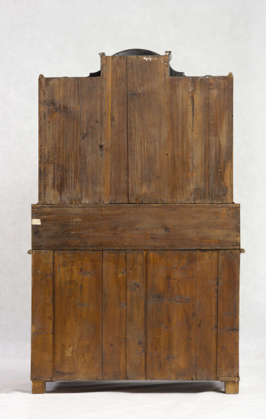 Sekretera trójkondygnacyjna. Podstawa w formie trzyszufladowej komody,wspartej na czterech kostkowych nogach. Środkowa część z prostokątnym skośnym, odchylnym blatem, obwiedziona profilowaną listwą, fronty szuflad wklęsło-wypukłe. Po bokach ukośnego blatu po dwie szufladki. Nastawa cofnięta z dwoma rzędami szufladek po bokach (po 4); część środkowa wyższa, w niej półkoliście wypukłe drzwiczki i dwie szufladki nad i pod drzwiczkami. Fronty szuflad faliste jak i wykrój profilowanego zwieńczenia. Powierzchnia intarsjowana, dekorowana fornirem: na drzwiczkach kompozycja w formie gałązki z kwiatami, na blacie symetrycznie ułożone dwa ornamenty liściasto-wolutowe,  na szufladach poziome prostokątny ujęte w ciemne ramki. Nakładane brązy:  szyldziki do kluczy, antabki i kuliste uchwyty. 
