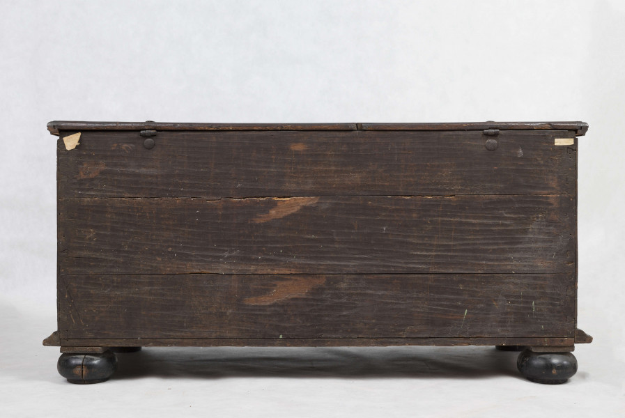 Korpus prostokątny, zamykany od góry gładkim  wiekiem na zawiasach, cztery nogi w kształcie silnie spłaszczonych kul. Ściana frontowa podzielona pionowo na dwie części, w których umieszczone są poziome płyciny w kształcie prostokąta o ściętych narożach wypełnionych trójkątnymi rautami, ujęte w profilowane ramy. Podziałowi odpowiadają w górnej części trzy rzeźbione maski lwów. Wzdłuż boków ściany frontowej i bocznych - lekko wgłębione płyciny w formie wydłużonych owali. Wszystkie płyciny i rauty obwiedzione ornamentem wody. W ścianach bocznych zamocowane masywne metalowe antaby.  Wewnątrz żelazny zamek nakładany.    