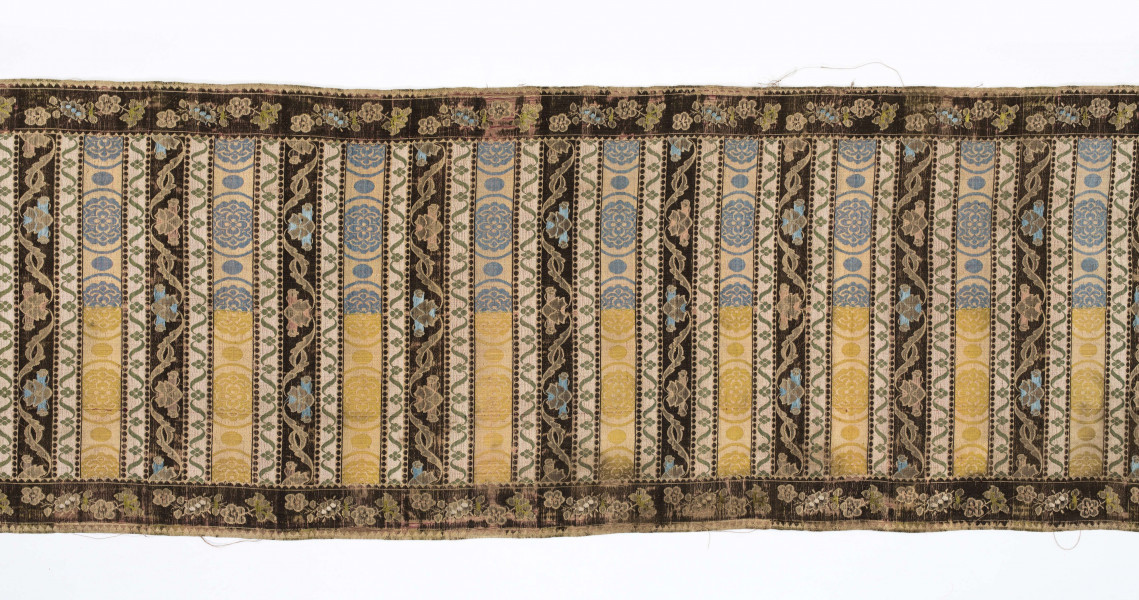 Pas 4-stronny. 
Ornament: głowy - krzaczki kwiatowe w wazonach; środek - poprzeczne pólka w układzie a,b,a,c (a - motyw wijacego sie wężyka, b - medaliony z rozetami, c - wić kwiatowa), obrzeża pionowe z gałązkami kwiatowymi, poziome - jak pólka c.
Koloryt: prawa strona - tło słomkowe, nieznacznie zróżnicowane w każdej połowie, zielen, błękit, żółty; lewa strona - tło brązowe głów i obrzeżeń, ornament - tło słomkowe, wzór j.w..
Fredzla doszyta jedwabna, pleciona, słomkowożółta