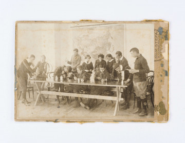 Na pierwszym planie ławka przy której 9 uczniów i 3 uczennice przeprowadzają doświadczenia. Na ławce widać przyrządy do doświadczeń chemicznych. W głębi sali na ścianie widać powieszoną mapę.