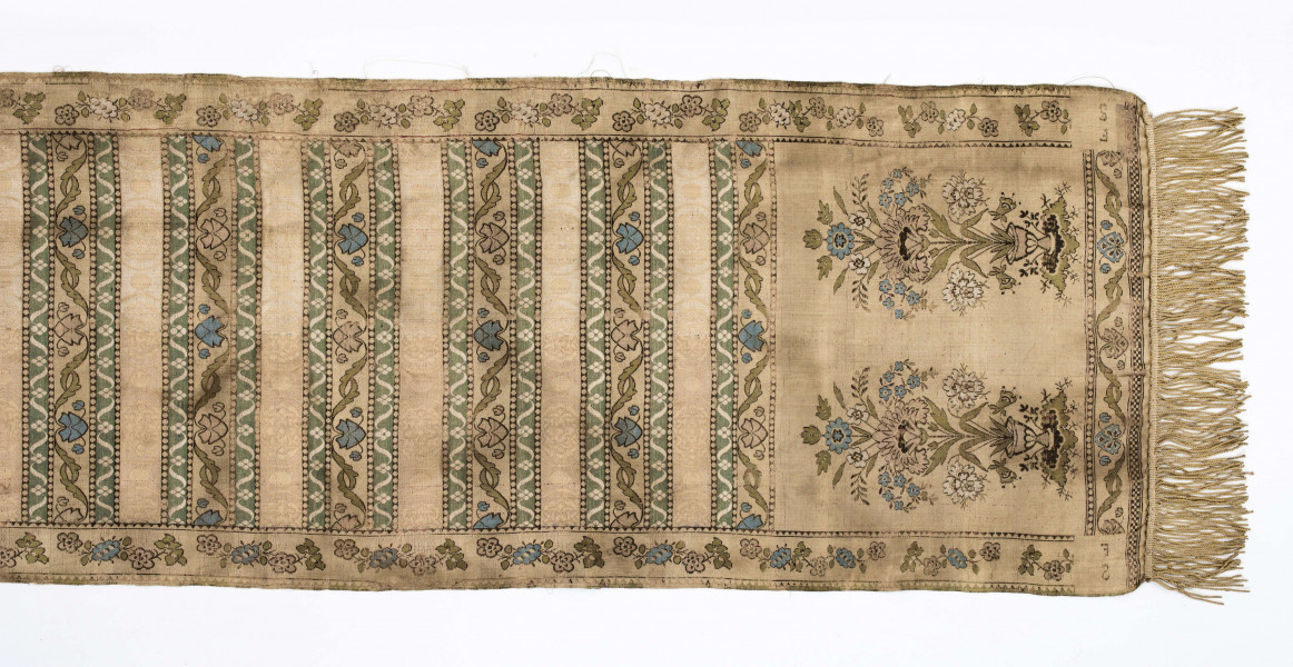 Pas 4-stronny. 
Ornament: głowy - krzaczki kwiatowe w wazonach; środek - poprzeczne pólka w układzie a,b,a,c (a - motyw wijacego sie wężyka, b - medaliony z rozetami, c - wić kwiatowa), obrzeża pionowe z gałązkami kwiatowymi, poziome - jak pólka c.
Koloryt: prawa strona - tło słomkowe, nieznacznie zróżnicowane w każdej połowie, zielen, błękit, żółty; lewa strona - tło brązowe głów i obrzeżeń, ornament - tło słomkowe, wzór j.w..
Fredzla doszyta jedwabna, pleciona, słomkowożółta