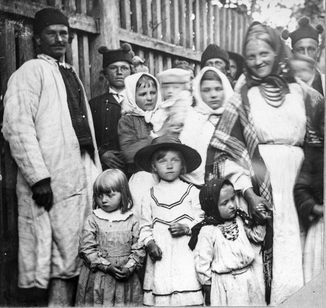 Fotografia czarno-biała przedstawia grupę dzieci i dorosłych w strojach ludowych na tle wysokiego drewnianego płotu. 