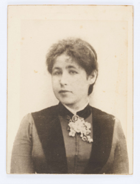 Głowa lekko skręcona w prawo, z opaską na głowie, w kamizelce, bluzka z broszką pod szyją.