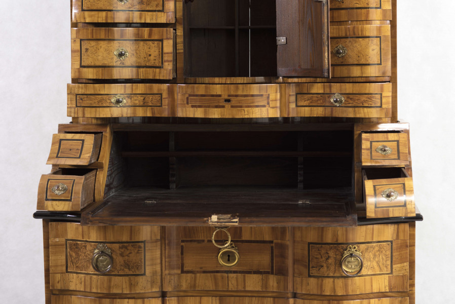 Sekretera trójkondygnacyjna. Podstawa w formie trzyszufladowej komody,wspartej na czterech kostkowych nogach. Środkowa część z prostokątnym skośnym, odchylnym blatem, obwiedziona profilowaną listwą, fronty szuflad wklęsło-wypukłe. Po bokach ukośnego blatu po dwie szufladki. Nastawa cofnięta z dwoma rzędami szufladek po bokach (po 4); część środkowa wyższa, w niej półkoliście wypukłe drzwiczki i dwie szufladki nad i pod drzwiczkami. Fronty szuflad faliste jak i wykrój profilowanego zwieńczenia. Powierzchnia intarsjowana, dekorowana fornirem: na drzwiczkach kompozycja w formie gałązki z kwiatami, na blacie symetrycznie ułożone dwa ornamenty liściasto-wolutowe,  na szufladach poziome prostokątny ujęte w ciemne ramki. Nakładane brązy:  szyldziki do kluczy, antabki i kuliste uchwyty. 
