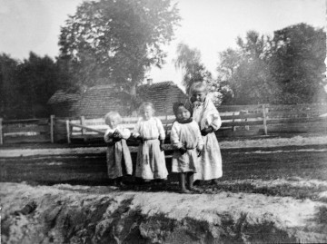 Fotografia czarno-biała przedstawia czworo małych dzieci w strojach ludowych, na tle drewnianego płotu i chaty wiejskiej otoczonej przez rozłożyste drzewa. 