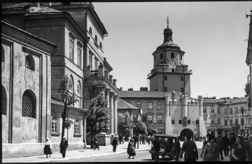 Fotografia czarnobiała z widokiem na plac Łokietka, Bramę Krakowską i fasadę Nowego Ratusza w Lublinie. Na pierwszym planie wylot ulicy Krakowskie Przedmieście z przechodniami na chodnikach i jednym zaparkowanym wzdłuż drogi autem widocznym od tyłu. Brama Krakowska otynkowana, zegar wskazujący godzinę 11.00, obok niego po lewej stronie tabliczka informująca o temperaturze: + 24. Kamienice po obu stronach Bramy z licznymi szyldami lokali gastronomicznych, usługowych i handlowych. W prawym dolnym rogu widoczna lampa w formie podwójnej ozdobnej pastorałki. Pośrodku zdjęcia narożnik piętrowej kamienicy ze sklepem meblowym (ob. nieistniejącej). Po lewej stronie fragment kościoła pw. Św. Ducha i fasada Nowego Ratusza z portykiem kolumnowym i schodami do wejścia. Na portyku balkon, na murowanych słupkach balustrady donice z kwiatami. Przed Ratuszem i sklepem meblowym kilkumetrowe drzewka liściaste. 